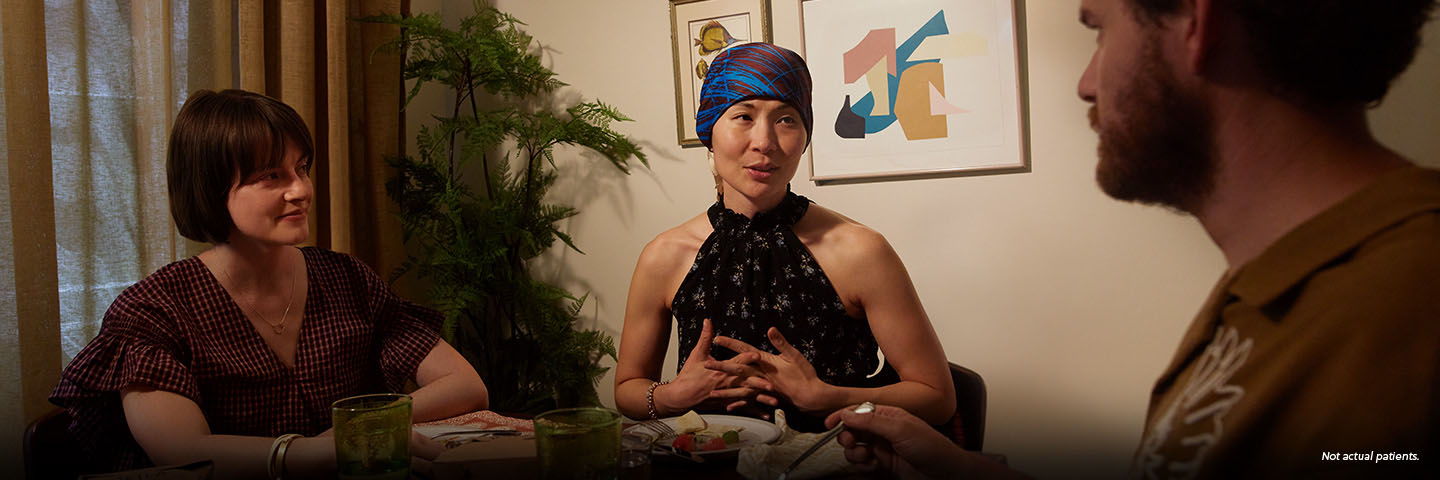 An Asian woman in her early 40s wearing a red and blue silk headscarf sits at a round dining table, smiling and talking with 2 friends, a white man and white woman of a similar age. They are finishing dinner, a takeout meal from one of their favorite restaurants. Not actual patients.