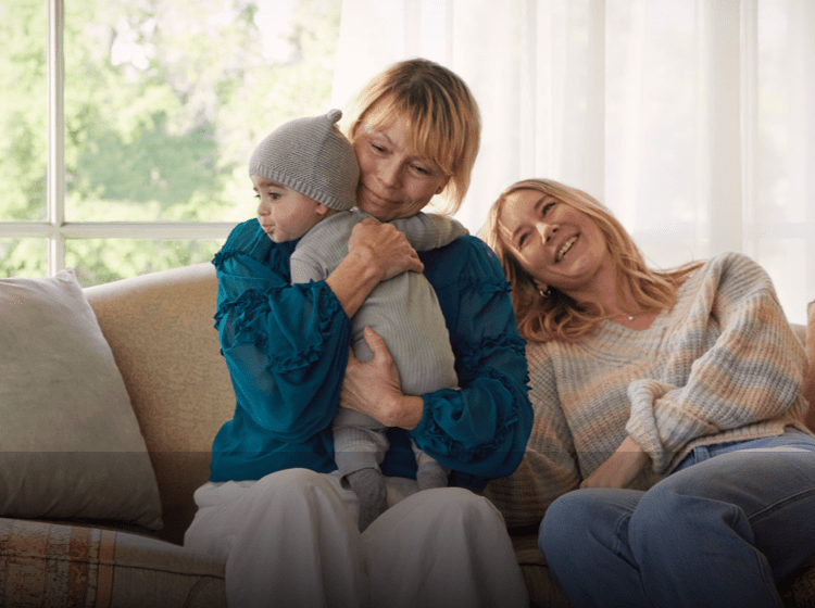 A White woman in her 60s with short blonde hair is sitting on the couch hugging her baby grandchild and smiling softly. Beside her, a White woman in her 30s rests her head on the couch and smiles. Actor portrayals.