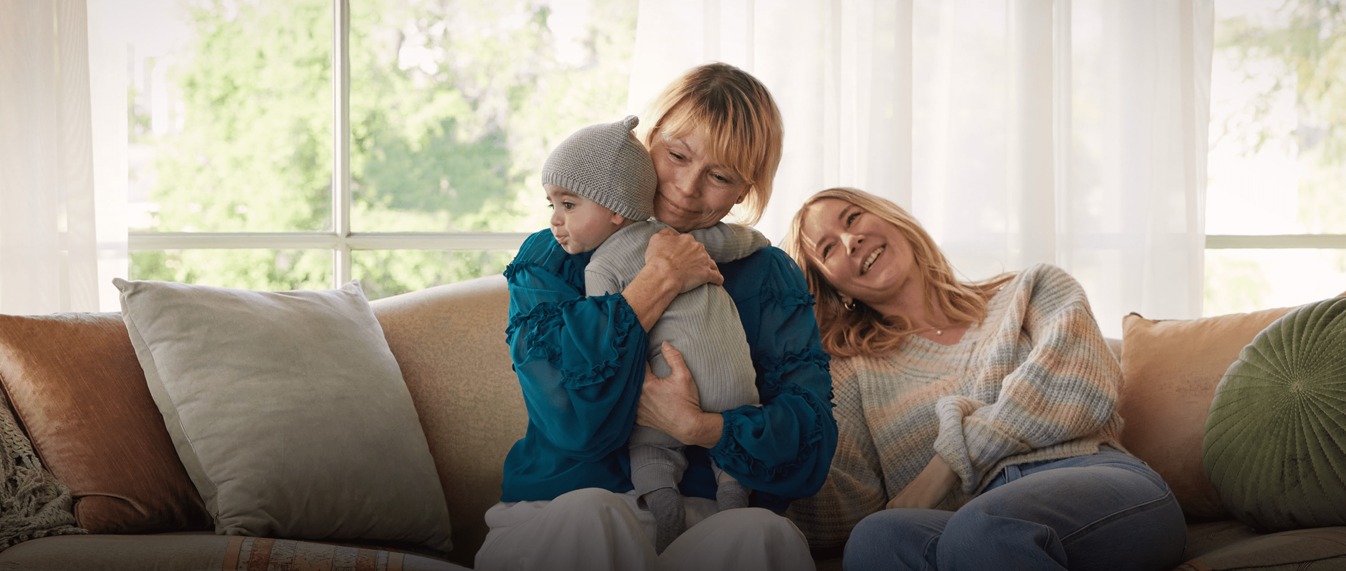 A White woman in her 60s with short blonde hair is sitting on the couch hugging her baby grandchild and smiling softly. Beside her, a White woman in her 30s rests her head on the couch and smiles. Actor portrayals.