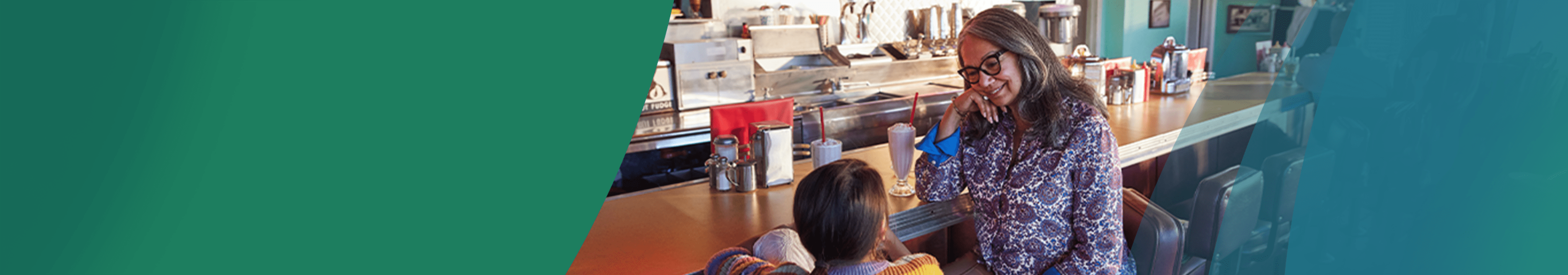 A Latina woman in her 60s and her young granddaughter are sitting on barstools at a vintage diner counter, looking at each other and smiling. The woman has long gray hair and bold glasses and is resting her elbow on the counter. Two strawberry milkshakes with whipped cream and long red straws are on the counter beside them. Actor portrayals.