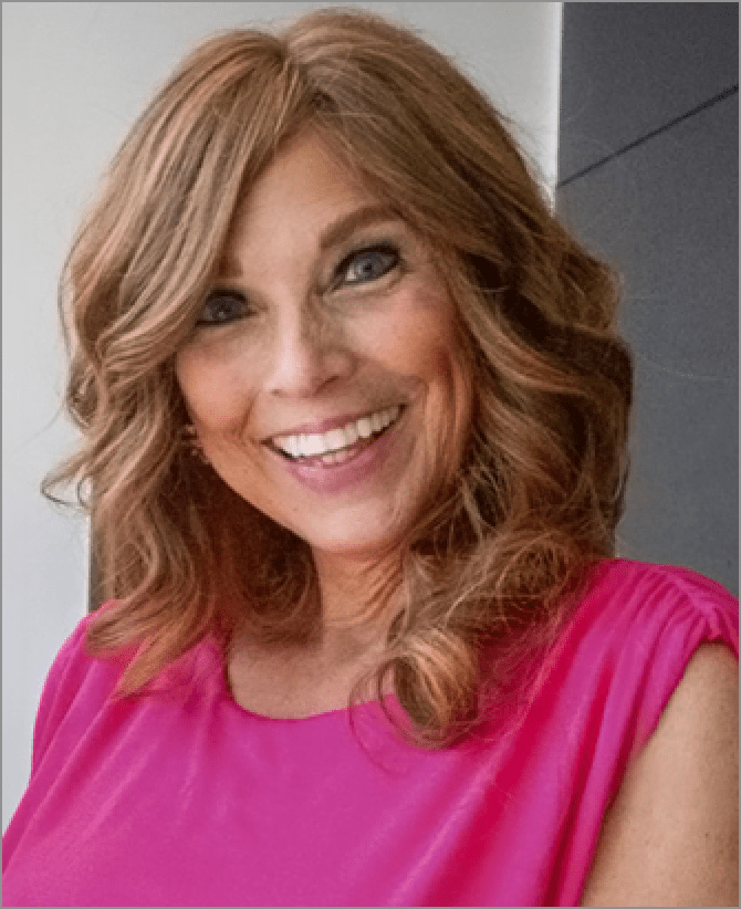 Jacquie, a woman receiving TRODELVY, is smiling at the camera. Jacquie is a White woman wearing a brown wig with blonde highlights, and a pink shirt.