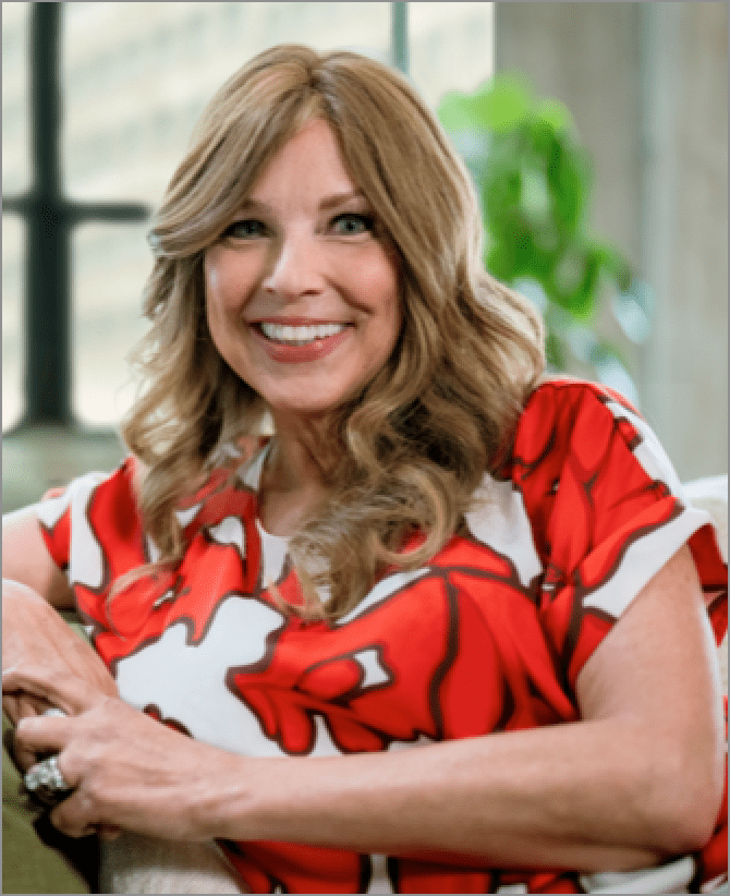Jacquie, a woman receiving TRODELVY, is sitting in a white armchair while smiling at the camera. Jacquie is a White woman wearing a brown wig with blonde highlights, and a shirt with a red and white pattern.