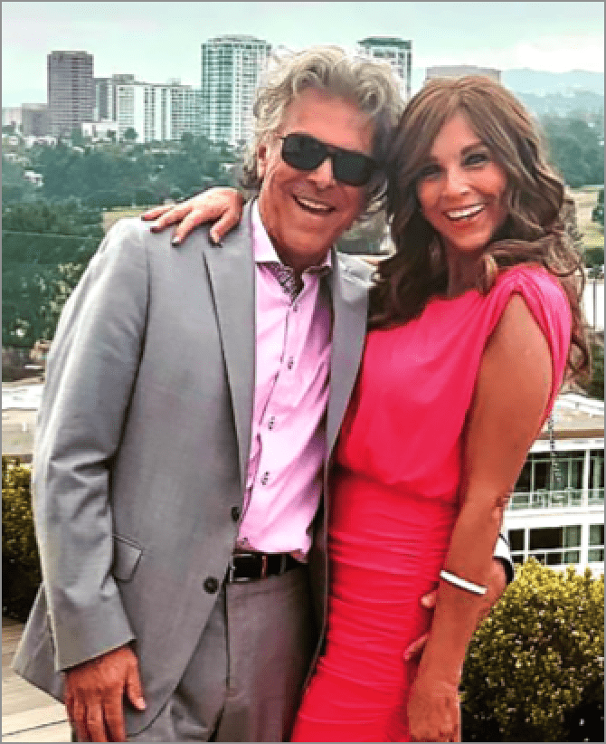 Jacquie, a woman receiving TRODELVY, is standing with her arm around her husband, Marc. Jacquie is a White woman, wearing a red dress and a wig. Marc is a White man with gray hair, wearing a gray suit and sunglasses.