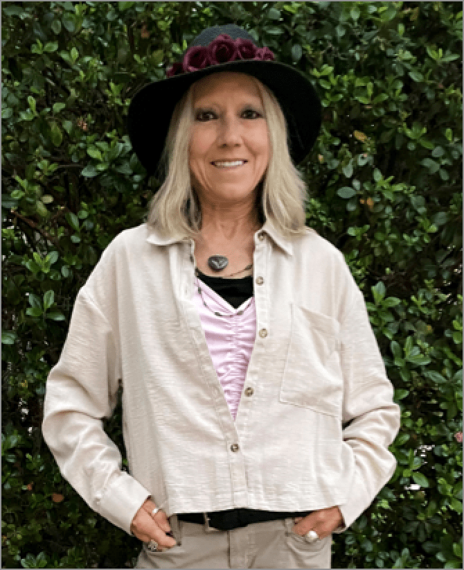 Karen, a woman receiving TRODELVY, is smiling at the camera. Karen is a White woman wearing a blonde wig, a black sun hat with roses on the brim, a tan jacket, a pink shirt, and brown pants.