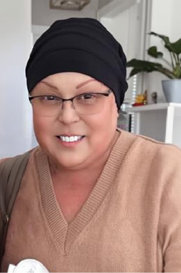 Lisa, a woman receiving TRODELVY, sits smiling at the camera. Lisa is a Latina woman wearing a headscarf, glasses, and a tan shirt.