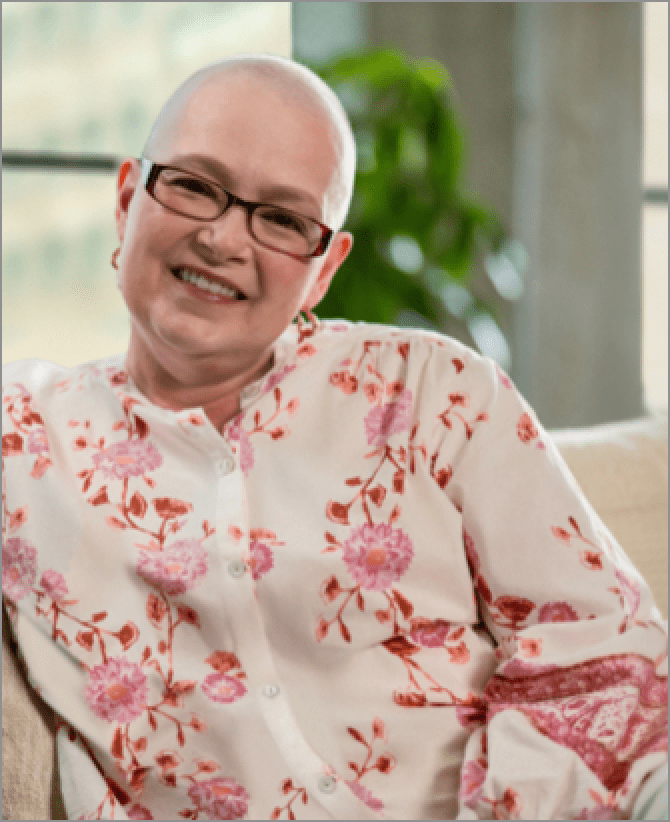 Tanya, a woman receiving TRODELVY, sits smiling at the camera. Tanya is a White woman with no hair, wearing glasses and a white shirt with a red floral pattern.