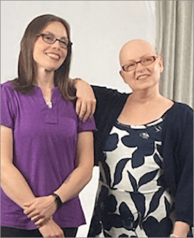 Tanya, a woman receiving TRODELVY, stands with her care partner, both smiling at the camera. Tanya is a White woman with no hair, wearing glasses and a black and white dress with a black jacket. Tanya's care partner is a White woman with brown hair, wearing glasses, a purple shirt, and black pants.