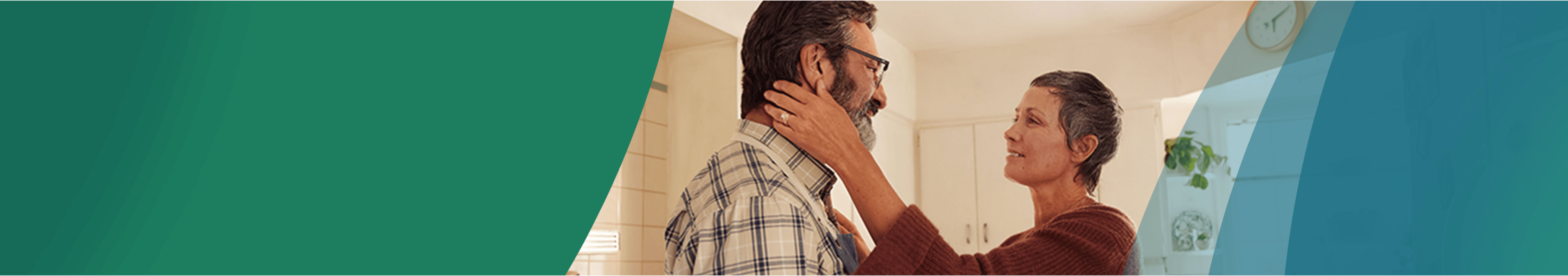 A White woman in her 50s with short gray hair is standing in the kitchen with her partner, a White man in his 50s. The woman is reaching out with both hands, one hand resting on the man's shoulder and the other gently holding the man's jaw. They are looking at each other fondly. Actor portrayals.