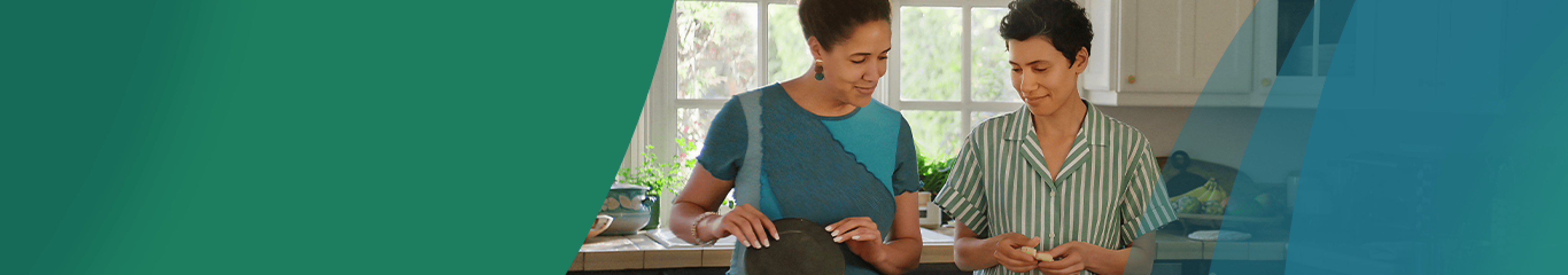 A Latina woman in her 40s with short, dark, curly hair stands in the kitchen with her partner, another Latina woman in her 40s. They are making tortillas from scratch, with one woman forming the masa and the other pressing it in the tortilla press. Actor portrayals.