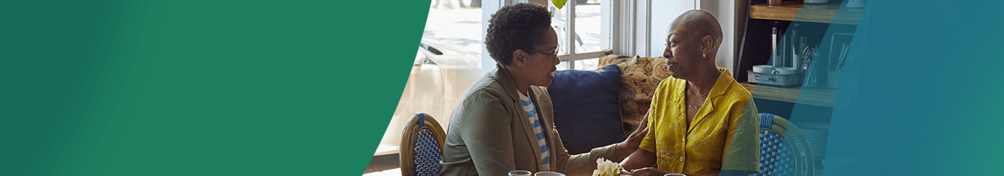 A Black woman in her 40s with closely shaved hair sits at a table in a coffee shop with her friend, another Black woman in her 40s. The friend is gently holding the other woman's arm in a gesture of support. Actor portrayals.
