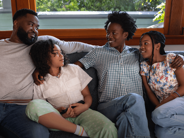 A Black woman in her 40s with short natural hair is cuddled on the living room couch with her family—a Black man in his 40s and their 2 preteen daughters. The woman has her arms around both of her daughters and is smiling at her husband. The family is close, leaning on each other and smiling. Actor portrayals.