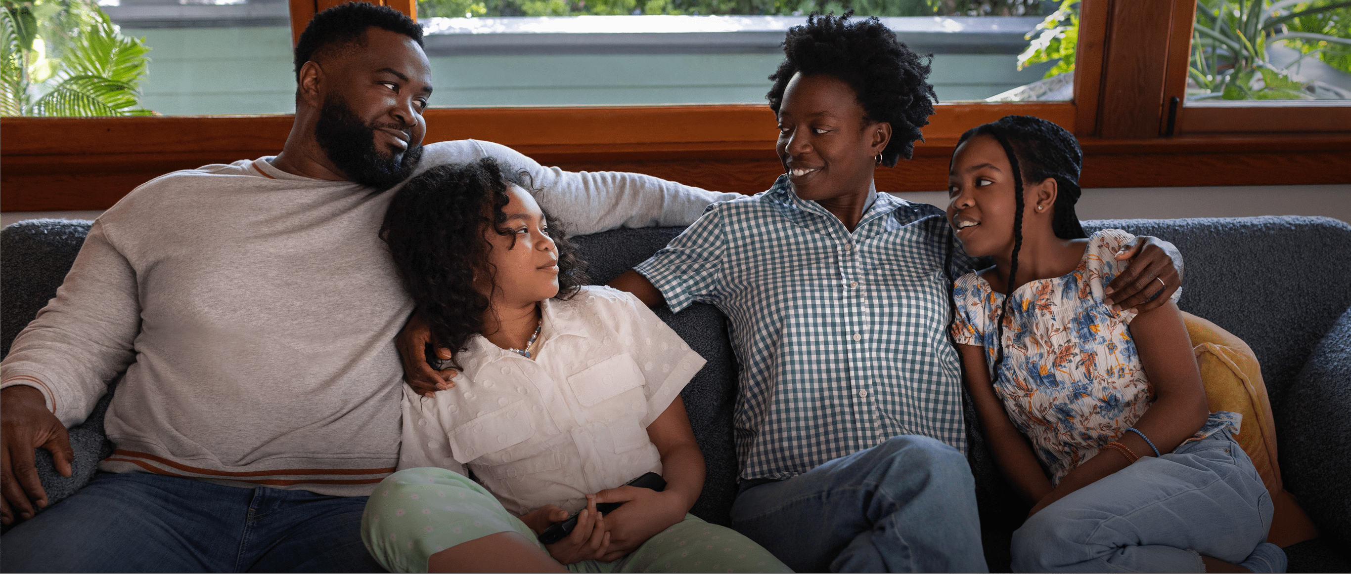 A Black woman in her 40s with short natural hair is cuddled on the living room couch with her family—a Black man in his 40s and their 2 preteen daughters. The woman has her arms around both of her daughters and is smiling at her husband. The family is close, leaning on each other and smiling. Actor portrayals.