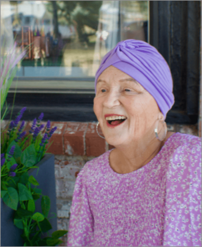 Jana, a woman receiving TRODELVY, sits smiling at the camera. Jana is a White woman wearing a purple headscarf, gold earrings, and a purple floral-patterned shirt.