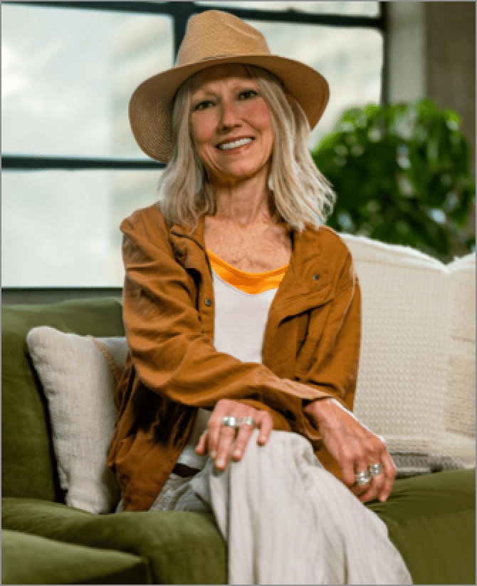 Karen, a woman receiving TRODELVY, is smiling at the camera. Karen is a White woman wearing a blonde wig, a brown sun hat, a brown leather jacket, and a white shirt and white pants.