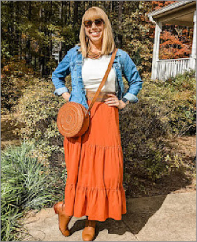 Katherine, a woman receiving TRODELVY, is standing outside smiling at the camera. Katherine is a White woman wearing a blonde wig, sunglasses, a denim jacket, a white shirt, an orange skirt, a leather purse, and brown leather boots.