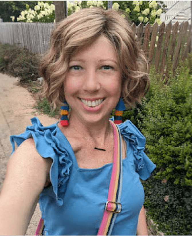 Katherine, a woman receiving TRODELVY, is standing outside and smiling at the camera. Katherine is a White woman wearing a blonde wig and a blue shirt.