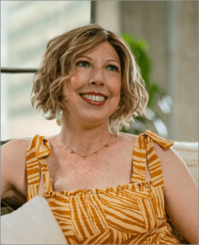 Katherine, a woman receiving TRODELVY, sits smiling at the camera. Katherine is a White woman wearing a blonde wig and a striped dress.
