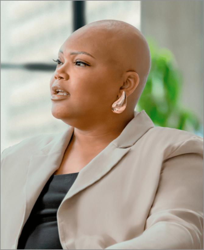 Tiana, a woman receiving TRODELVY, is sitting on a couch. Tiana is a Black woman with no hair, wearing a black shirt under a tan jacket, and denim jeans.