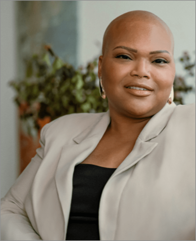 Tiana, a woman receiving TRODELVY, is sitting on a couch while smiling at the camera. Tiana is a Black woman with no hair, wearing gold earrings, and a black shirt under a tan jacket.