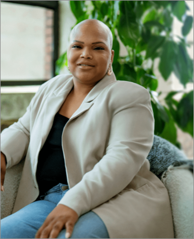 Tiana, a woman receiving TRODELVY, is sitting on a couch while smiling at the camera. Tiana is a Black woman with no hair, wearing a black shirt under a tan jacket, and denim jeans.