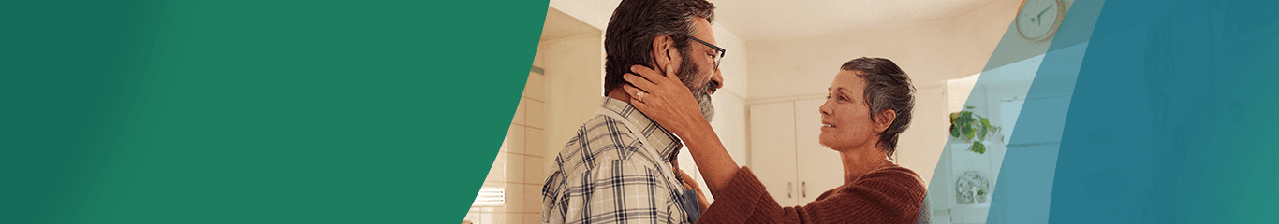 A Latina woman in her 50s with short gray hair is standing in the kitchen with her partner, a Latino man in his 50s. The woman is reaching out, with one hand resting on the man's shoulder and the other hand gently holding the man's jaw. They are looking at each other fondly. Actor portrayals.
