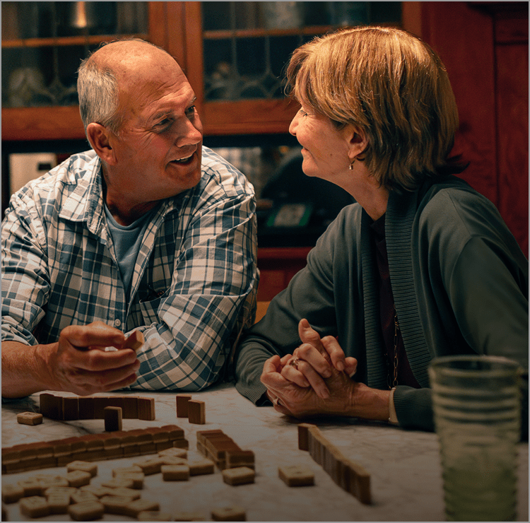 A White man, a White woman, and an Asian woman in their 60s are sitting at a dining table, laughing as they play a board game. Actor portrayals.