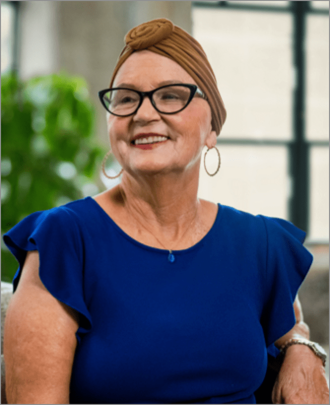 Jana, a woman receiving TRODELVY, sits smiling on a couch. Jana is a White woman wearing a headscarf, glasses, hoop earrings, and a blue shirt.