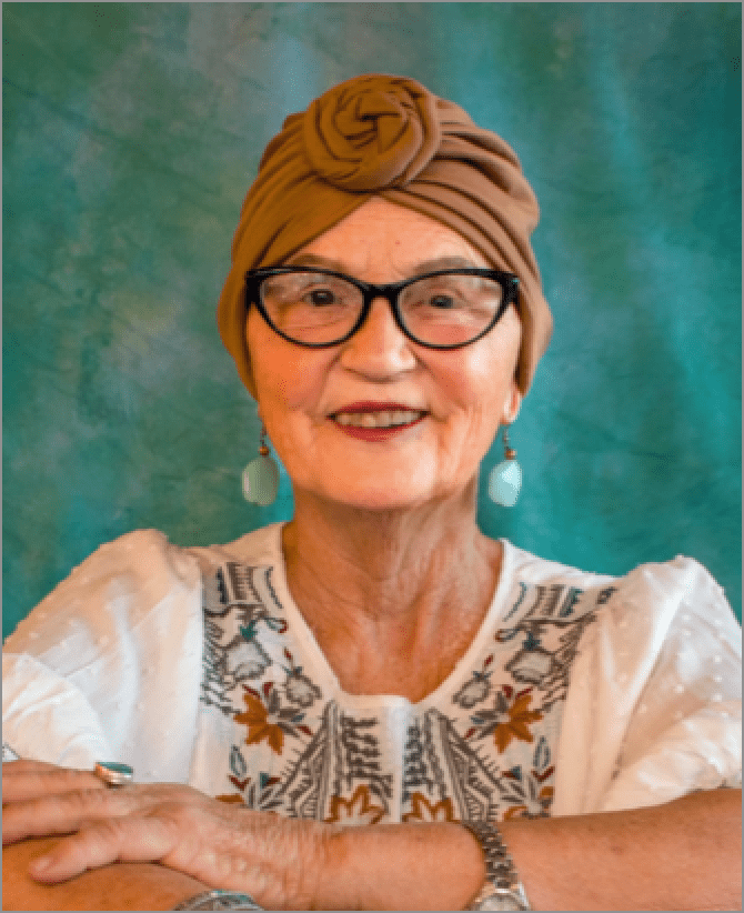 Jana, a woman receiving TRODELVY, sits smiling at the camera. Jana is a White woman wearing a headscarf, glasses, and a white patterned shirt.