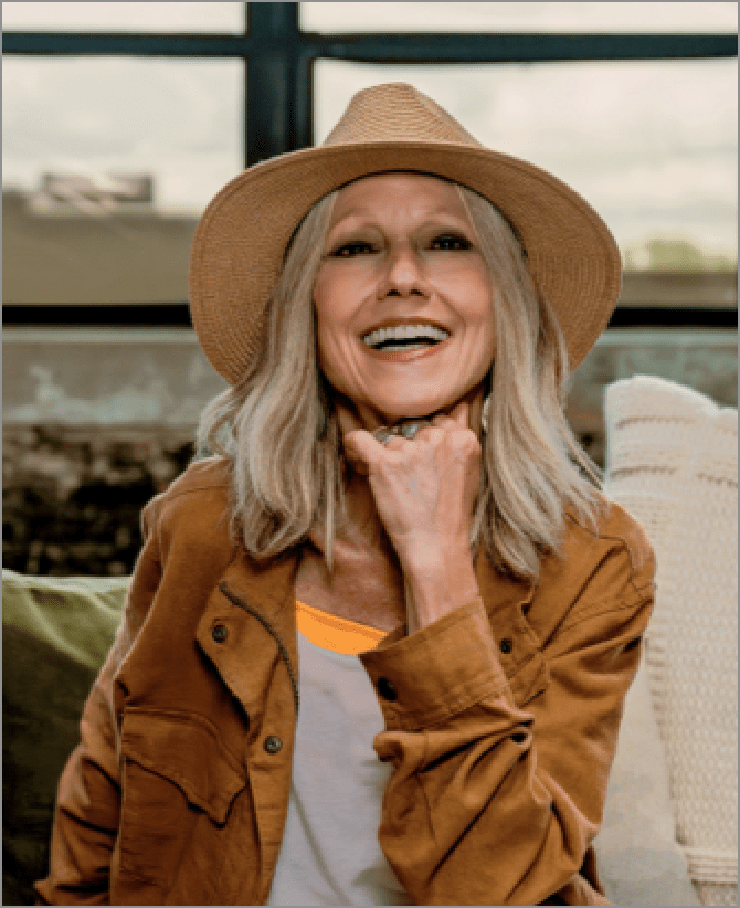 Karen, a woman receiving TRODELVY, is smiling at the camera. Karen is a White woman wearing a blonde wig, a brown sun hat, a brown leather jacket, and a white shirt and white pants.