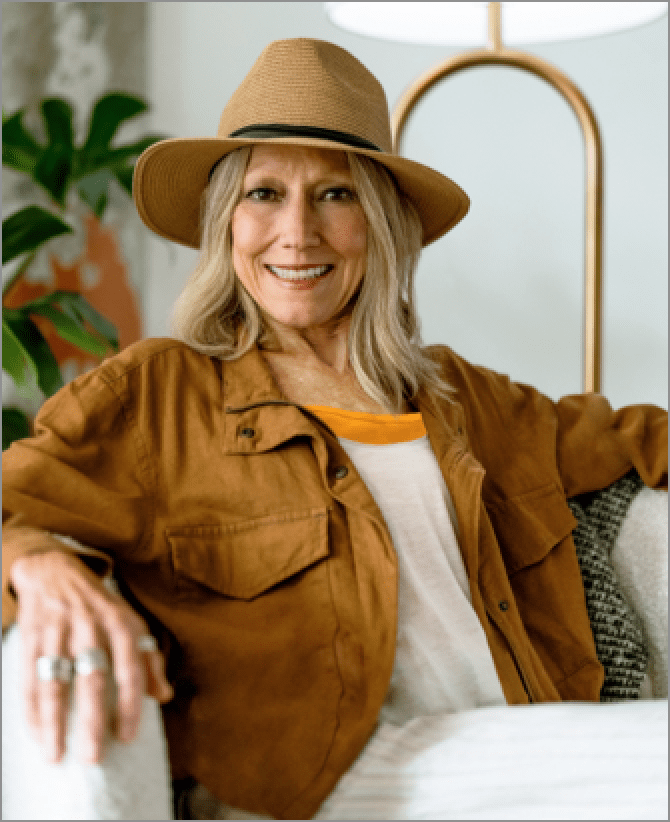 Karen, a woman receiving TRODELVY, is smiling at the camera. Karen is a White woman wearing a blonde wig, a brown sun hat, a brown leather jacket, and a white shirt and white pants.