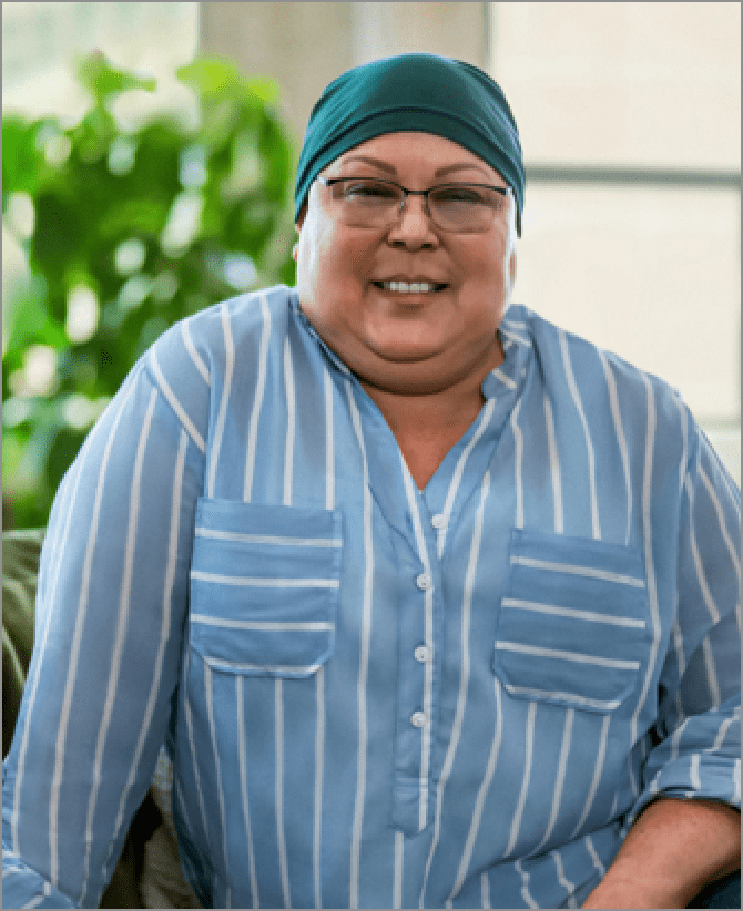 Lisa, a woman receiving TRODELVY, sits smiling at the camera. Lisa is a Latina woman wearing a headscarf, glasses, and a blue shirt with white stripes.