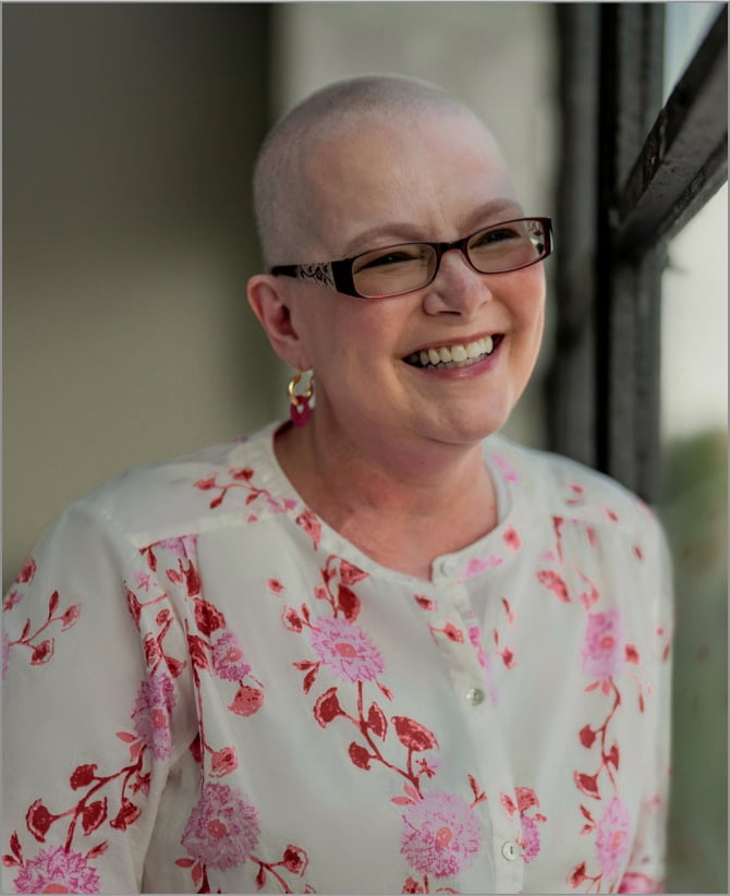 Tanya, a woman receiving TRODELVY, sits smiling at the camera. Tanya is a White woman with no hair, wearing glasses and a white shirt with a red floral pattern.