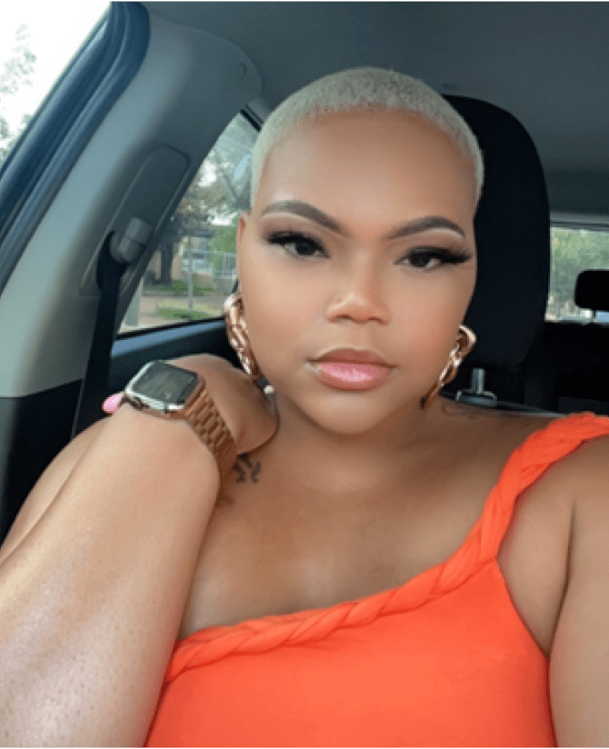 Tiana, a woman receiving TRODELVY, sits in her car smiling at the camera. Tiana is a Black woman with short blonde hair wearing an orange shirt, gold earrings, and a gold watch.