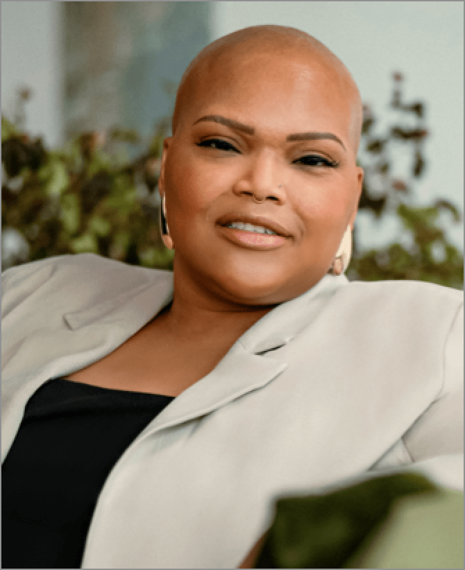 Tiana, a woman receiving TRODELVY, is sitting on a couch while smiling at the camera. Tiana is a Black woman with no hair, wearing gold earrings, and a black shirt under a tan jacket.