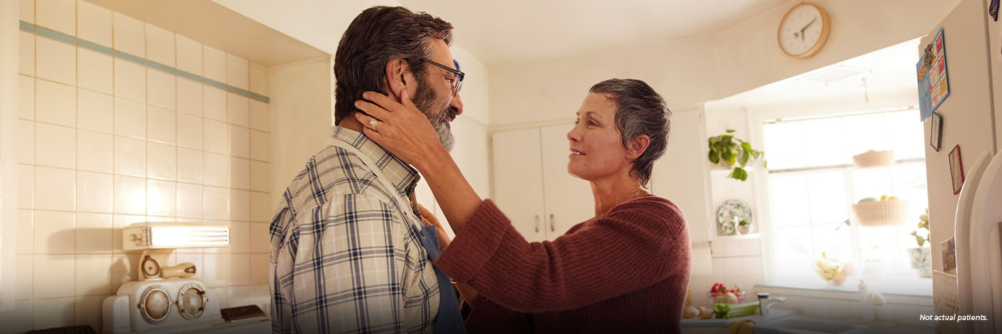 A Latina woman in her 50s with short gray hair is standing in the kitchen with her partner, a Latino man in his 50s. The woman is reaching out, with one hand resting on the man's shoulder and the other hand gently holding the man's jaw. They are looking at each other fondly. Not actual patients.