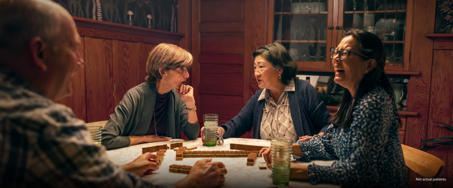 Two asian woman, a white man, and a white woman in their 60s are sitting at a dining room table, playing a board game and laughing. Not actual patients.