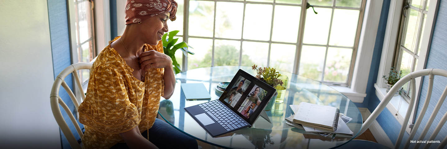 A Black woman in her early 40s wearing a paisley headscarf and a patterned yellow shirt sits in a brightly lit room at a round glass table smiling at her tablet screen, on a video call with her close group of friends. A potted plant, notebook, and notepad sit on the table with her tablet. Not actual patients.