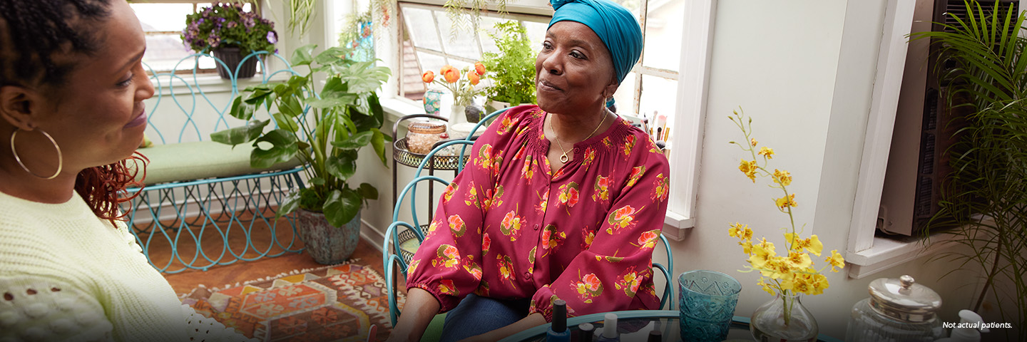 A Black woman in her mid-50s wearing a teal headscarf is sitting at a glass table in a brightly lit sunroom. She is with her friend, a Black woman in her late 40s with long braids and hoop earrings who is giving her a manicure. Colorful nail polish bottles are on the table. Not actual patients.