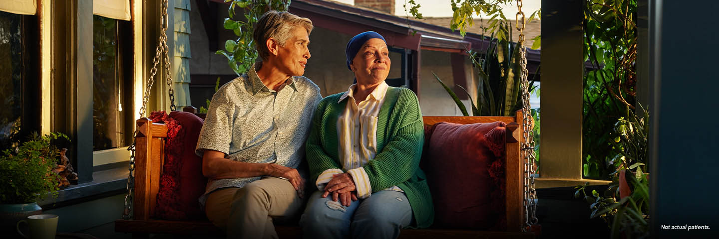 A white woman in her late 50s wearing a dark blue headscarf and a green sweater is sitting outside on a porch swing at sunset. Her partner, a white woman in their 50s with short gray hair, sits beside her with their arm across the back of the swing. Both are looking out into the yard, smiling. Not actual patients.