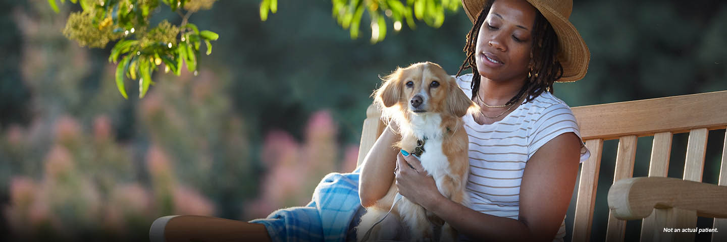 A Black woman in her 40s is sitting on a park bench outside, petting her small tan dog on her lap. The woman has chin-length twists and is wearing a sun hat. Not an actual patient.
