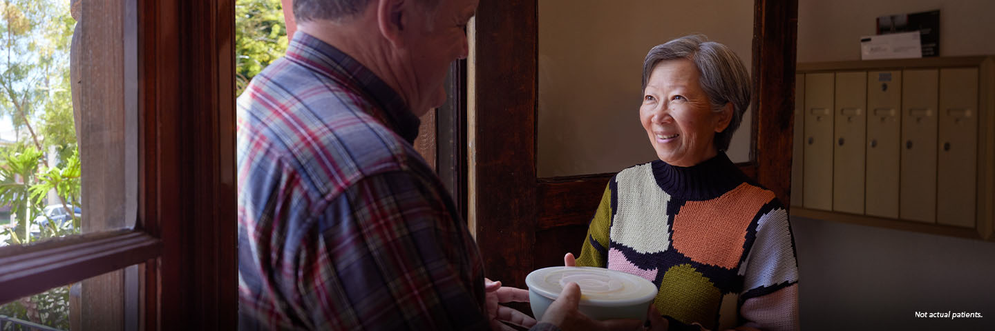 An Asian woman in her mid-60s with short gray hair meets her neighbor, a white man in his 60s, in the lobby of her apartment building. The man is handing her a container full of soup and they are smiling at each other. Not actual patients.