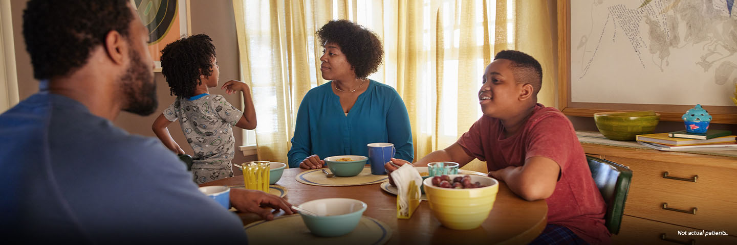 A Black woman in her mid-40s with short natural hair is sitting at her kitchen table at breakfast time with her partner, a Black man in his mid-40s, and 2 sons, aged 4 and 13. Both boys are in their pajamas, and the younger boy is kneeling up on his chair, talking excitedly. The woman is looking at him and smiling. Not actual patients.