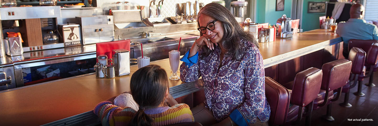 A Latina woman in her 60s and her young granddaughter are sitting on barstools at a vintage diner counter, looking at each other and smiling. The woman has long gray hair and bold glasses and is resting her elbow on the counter. Two strawberry milkshakes with whipped cream and long red straws are on the counter beside them. Not actual patients.