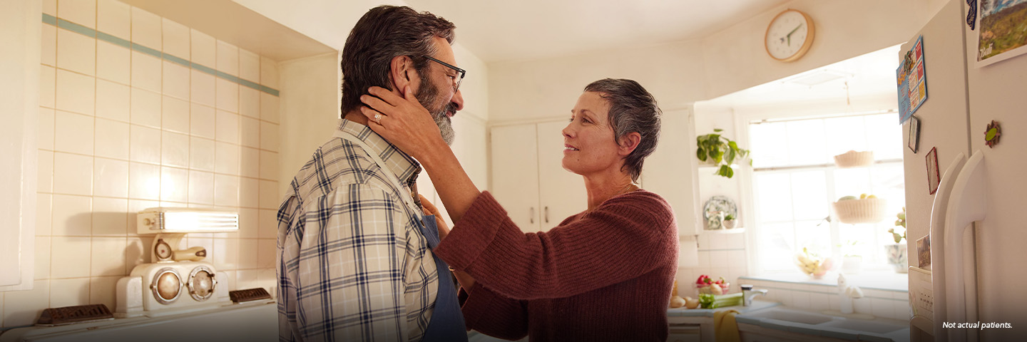 A white woman in her 50s with short gray hair is standing in the kitchen with her partner, a white man in his 50s. The woman is reaching out, with one hand resting on the man's shoulder and the other hand gently holding the man's jaw. They are looking at each other fondly. Not actual patients.