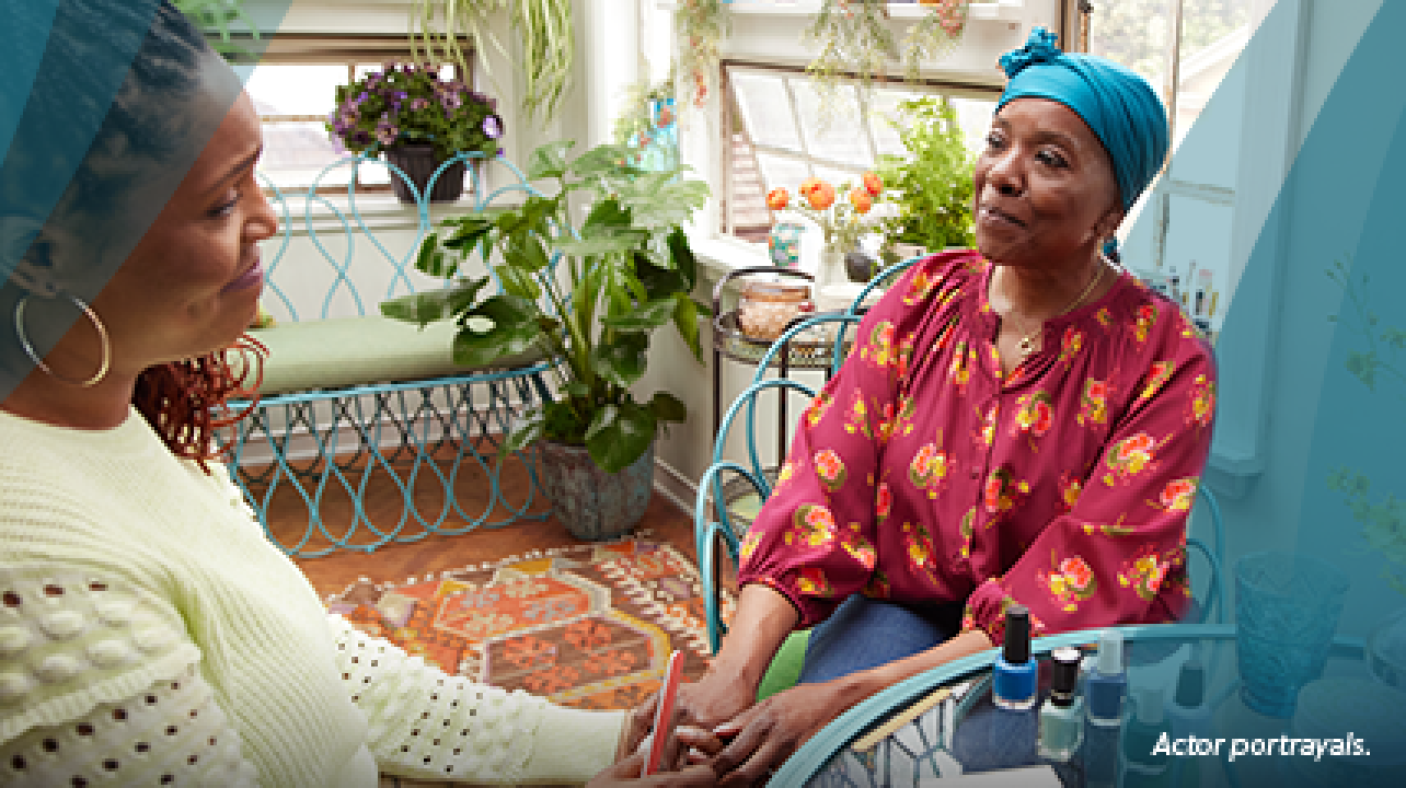 A Black woman in her mid-50s wearing a teal headscarf is sitting at a glass table in a brightly lit sunroom. She is with her friend, a Black woman in her late 40s with long braids and hoop earrings who is giving her a manicure. Colorful nail polish bottles are on the table. Actor portrayals.