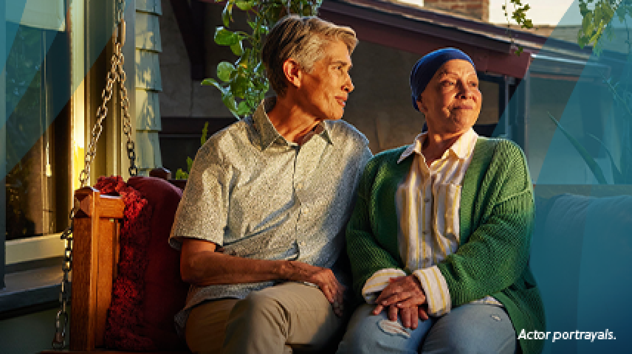 A white woman in her late 50s wearing a dark blue headscarf and a green sweater is sitting outside on a porch swing at sunset. Her partner, a white woman in their 50s with short gray hair, sits beside her with their arm across the back of the swing. Both are looking out into the yard, smiling. Actor portrayals.