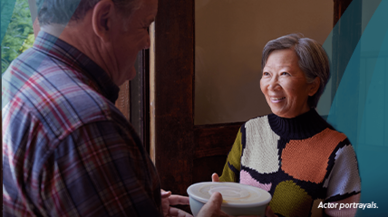 An Asian woman in her mid-60s with short gray hair meets her neighbor, a white man in his 60s, in the lobby of her apartment building. The man is handing her a container full of soup and they are smiling at each other. Actor portrayals.