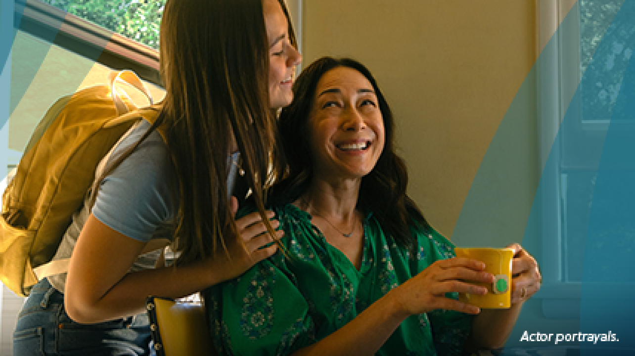 An Asian woman in her 50s is sitting at the kitchen table with a cup of tea and a slice of toast. Her teenage daughter is giving her a quick hug from behind as she is about to leave for school. They are looking at each other and smiling. Actor portrayals.