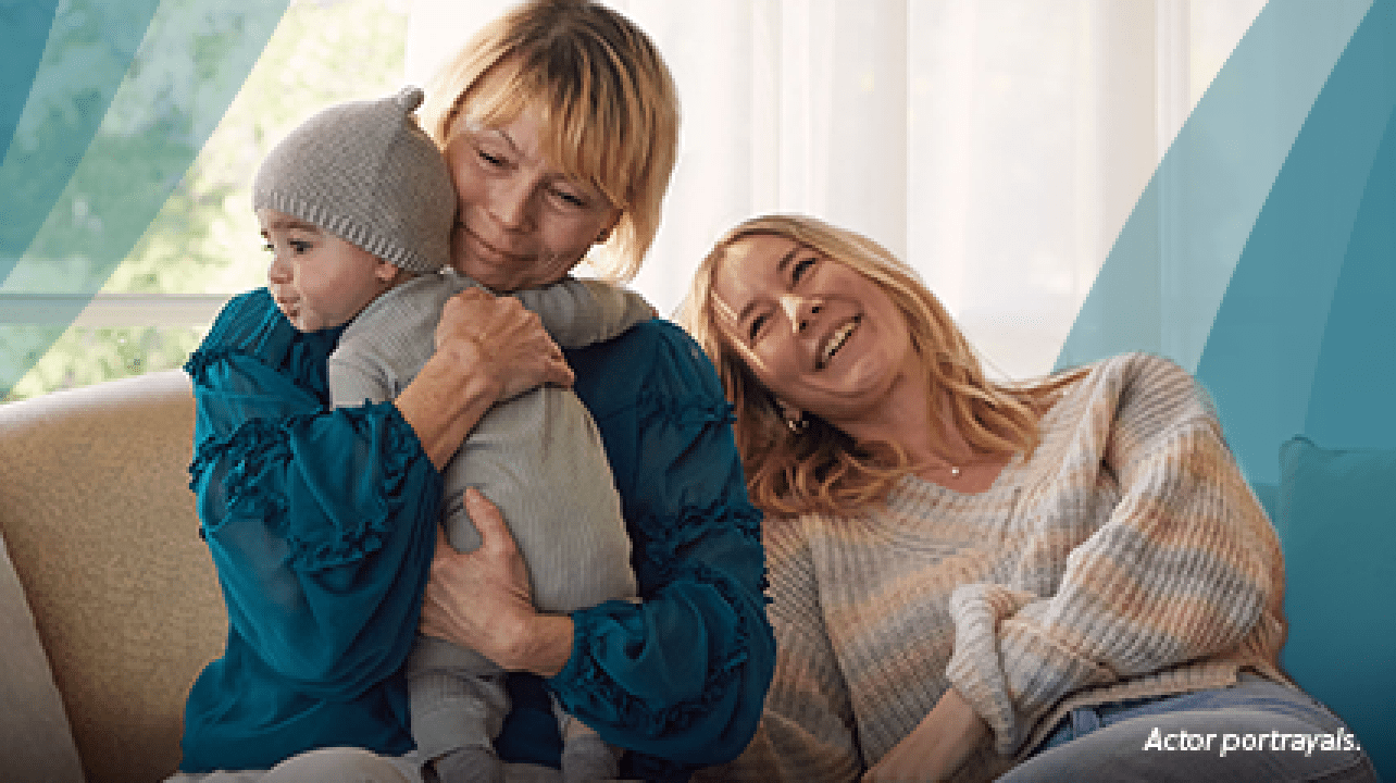 A white woman in her mid-60s with short blond hair is sitting on the couch hugging a baby tightly and smiling softly. Beside her, a white woman in her 30s rests her head on the couch and smiles. Actor portrayals.