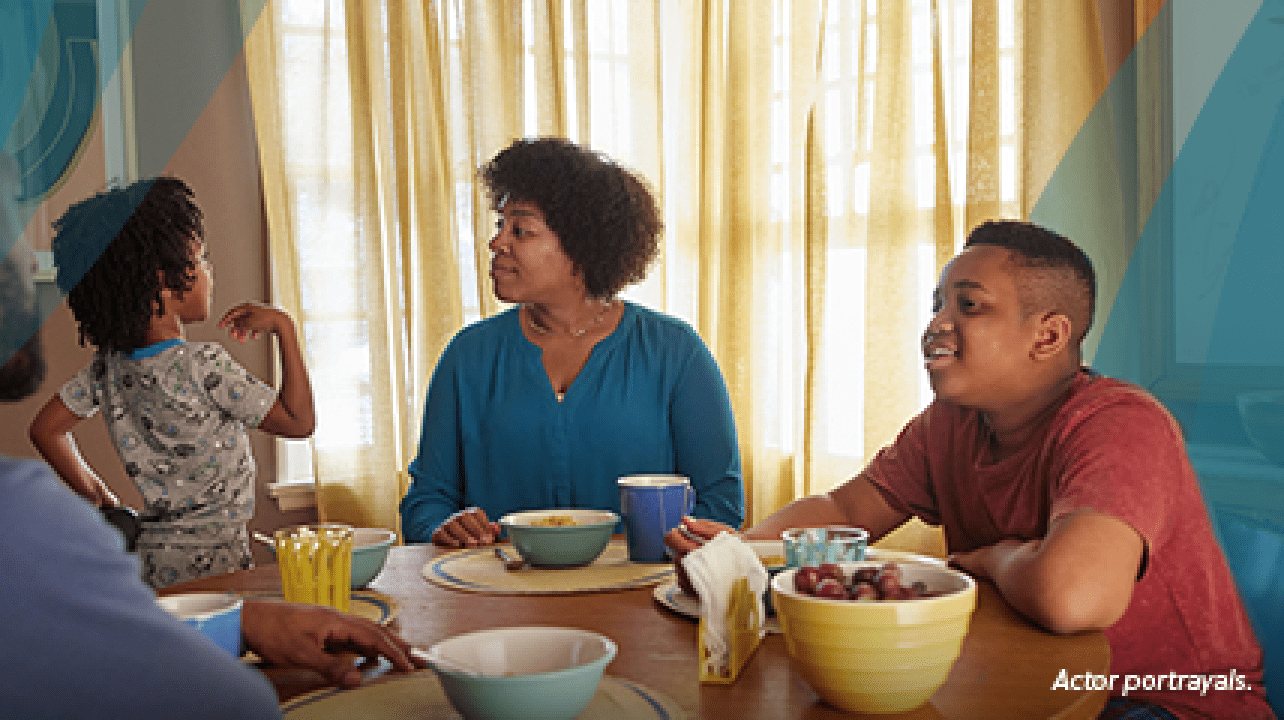 A Black woman in her mid-40s with short natural hair is sitting at her kitchen table at breakfast time with her partner, a Black man in his mid-40s, and 2 sons, aged 4 and 13. Both boys are in their pajamas, and the younger boy is kneeling up on his chair, talking excitedly. The woman is looking at him and smiling. Actor portrayals.